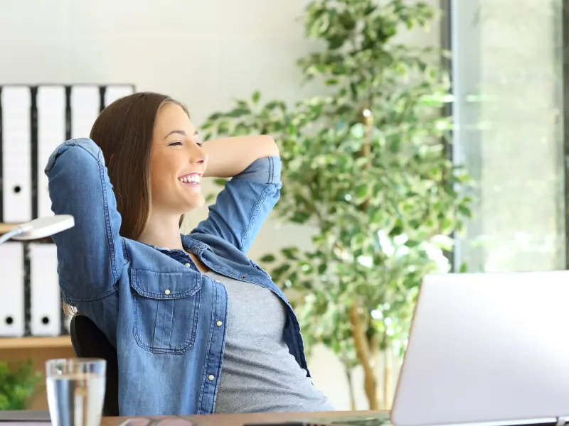 Gesundheitsförderung im Homeoffice. Eine Frau im Homeoffice, die lächelt und sich entspannt mit einer Tasse Tee am Schreibtisch sitzt.